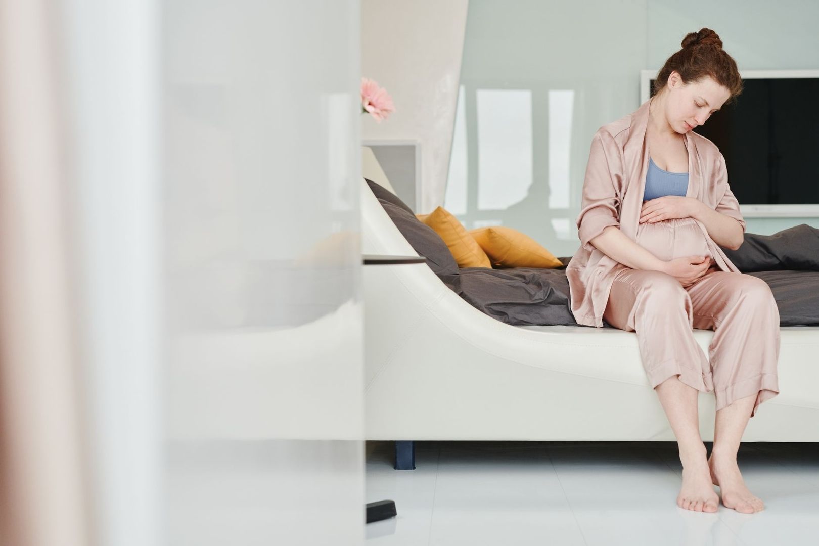 Gravid kvinna sitter på säng och håller händerna över bäckenet