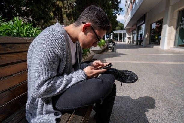 Ung person sitter framåtlutad och använder en smartphone på en bänk